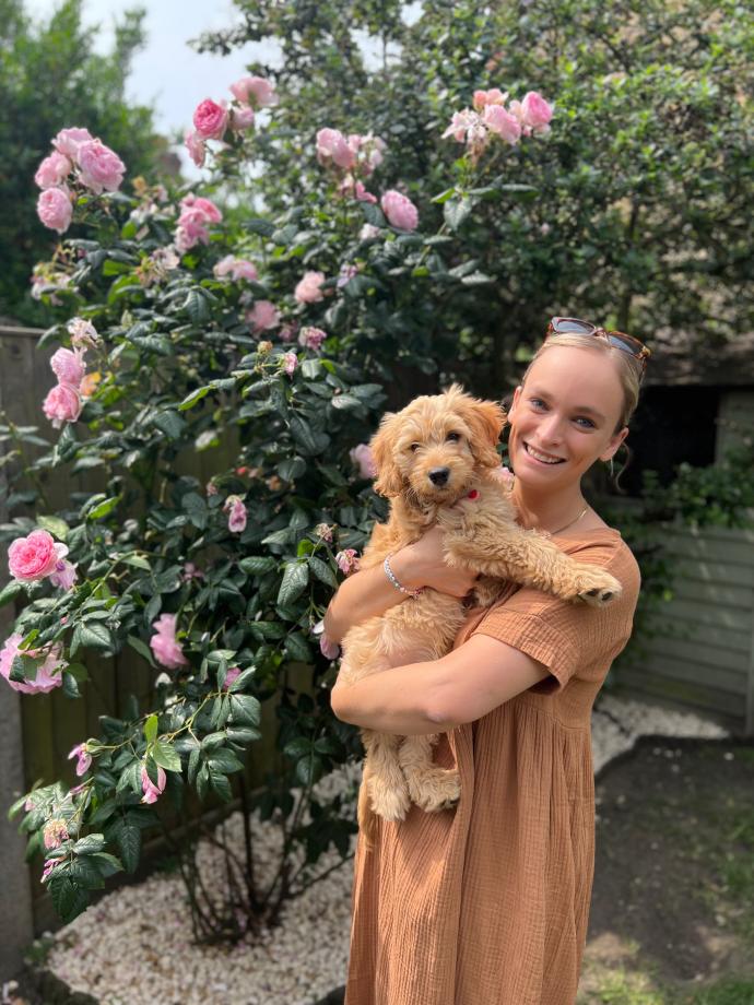 A photograph of Tannagh smiling, standing next to a rose bush and holding a puppy