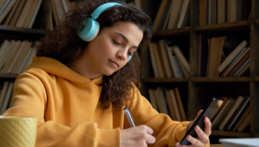 A teenage girl wearing a yellow hoodie and blue headphones studying in a library 